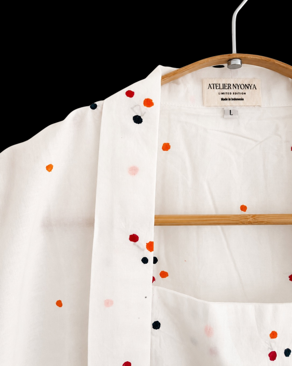 White open-front kimono-style top with colorful embroidered dots in orange, red, black, and pink, hanging on a wooden hanger against a black background. The neck label reads ATELIER NYONYA.