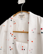 A white open-front kimono-style top with colorful embroidered dots, hanging on a wooden hanger against a black background. The brand label reads Atelier Nova.
