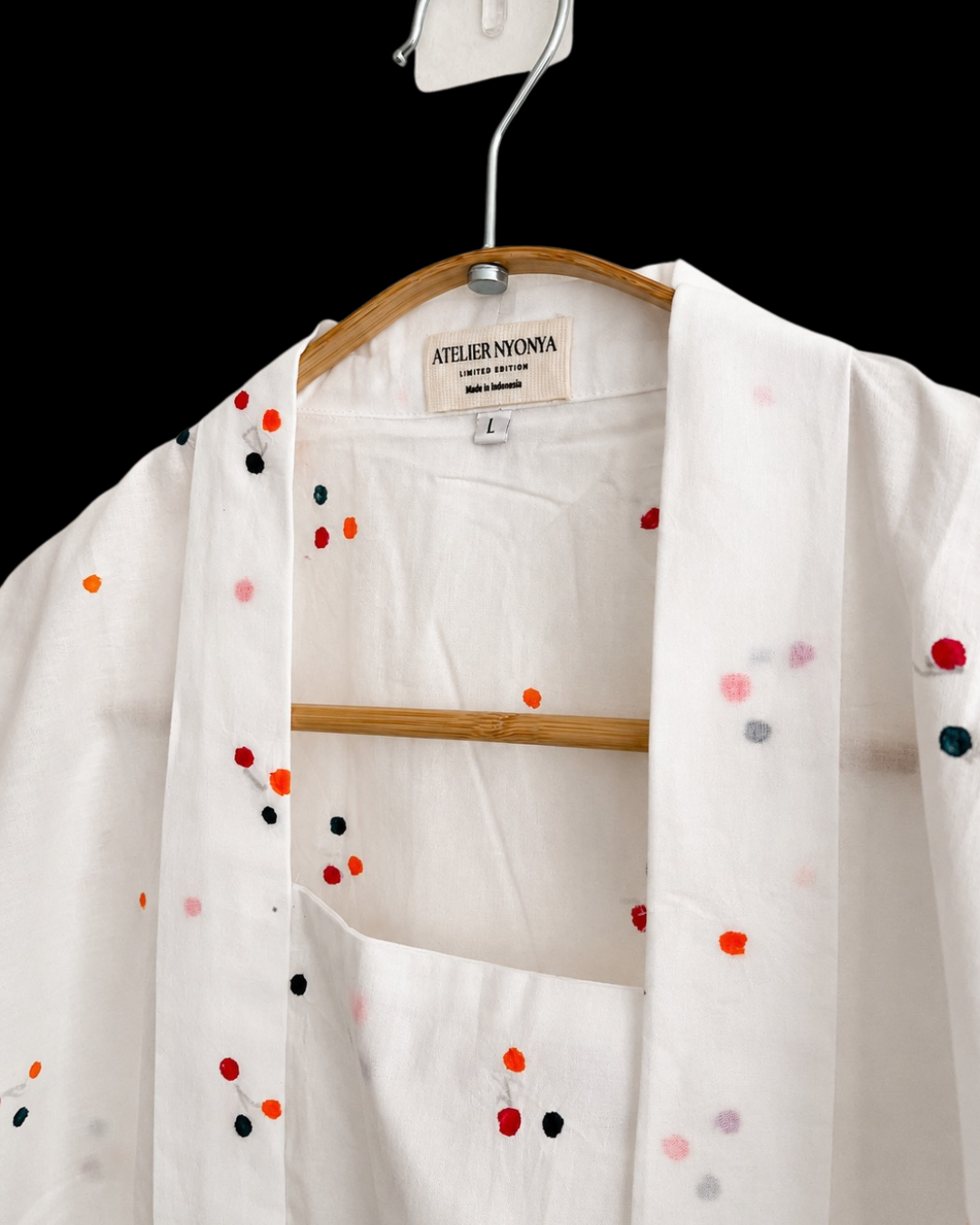 A white open-front kimono-style top with colorful embroidered dots, hanging on a wooden hanger against a black background. The brand label reads Atelier Nova.